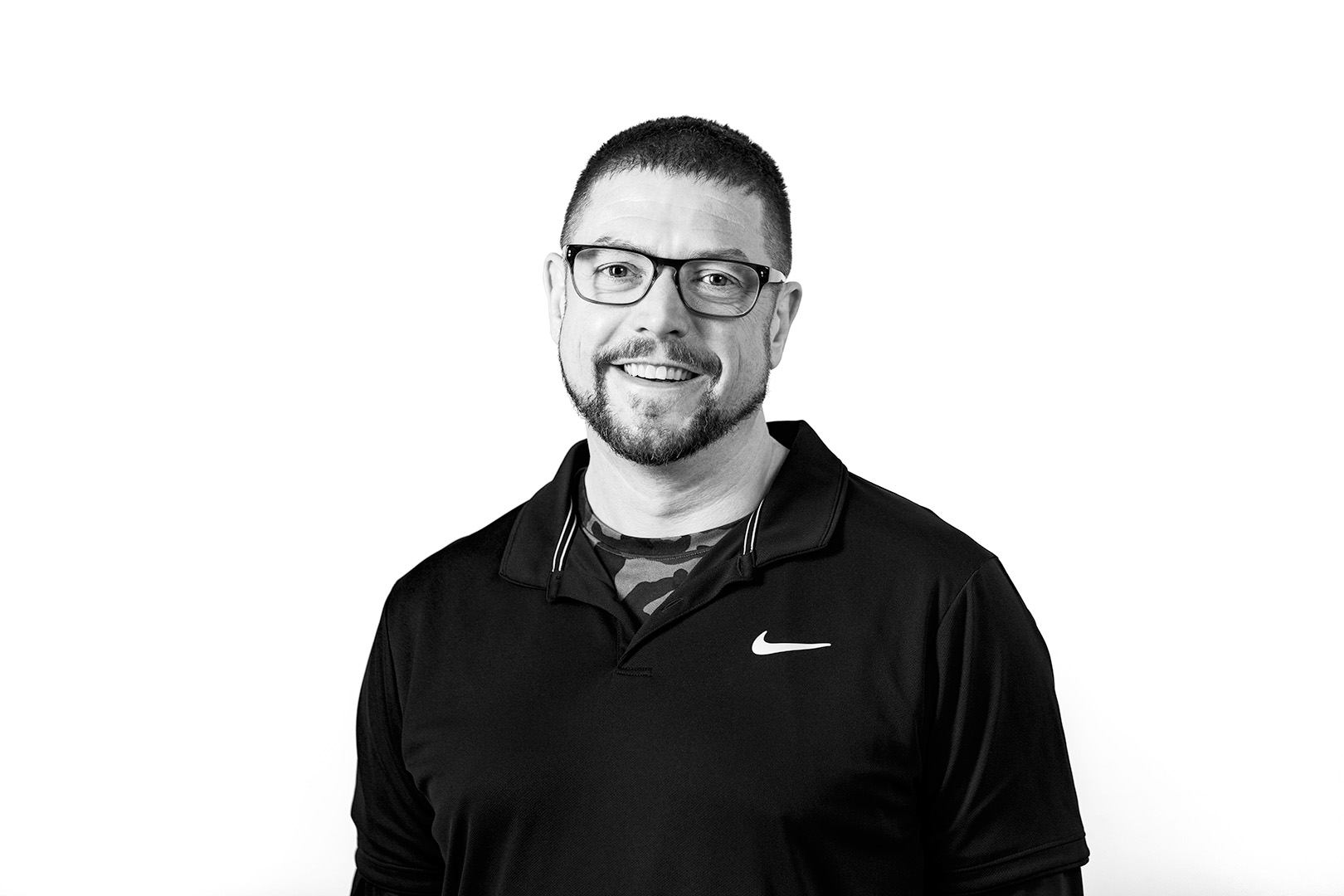 Portrait of Stew Smith: a black-and-white headshot of Stew smiling warmly. He has short brown hair, a close trimmed beard and rectangular glasses, and wears a black polo shirt, set against a plain white background.